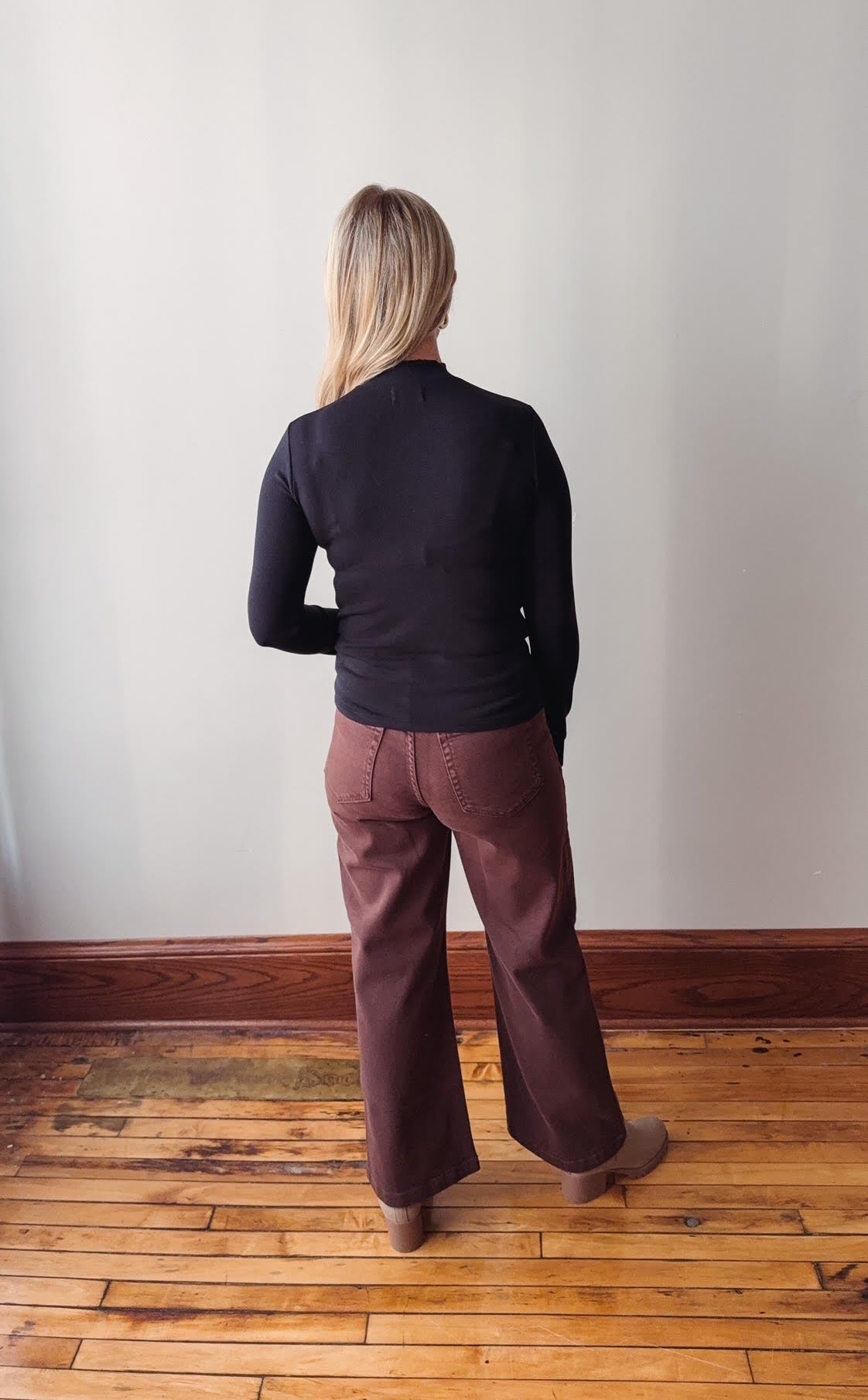 Person wearing a black sweater and brown pants standing against a white wall on a wooden floor.