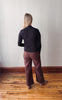 Person wearing a black sweater and brown pants standing against a white wall on a wooden floor.