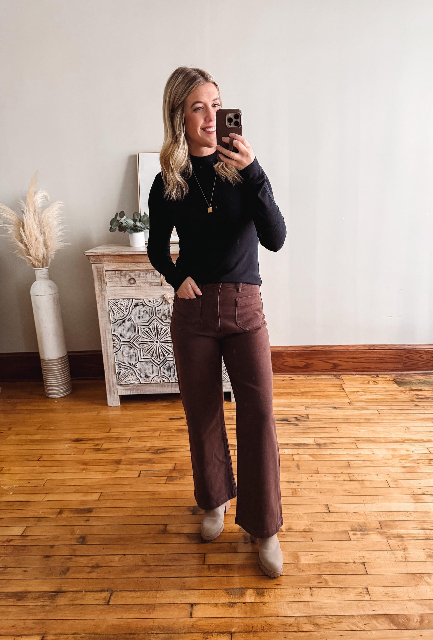 Woman taking a mirror selfie wearing a black top and brown pants in a room with wooden flooring and a decorative cabinet.