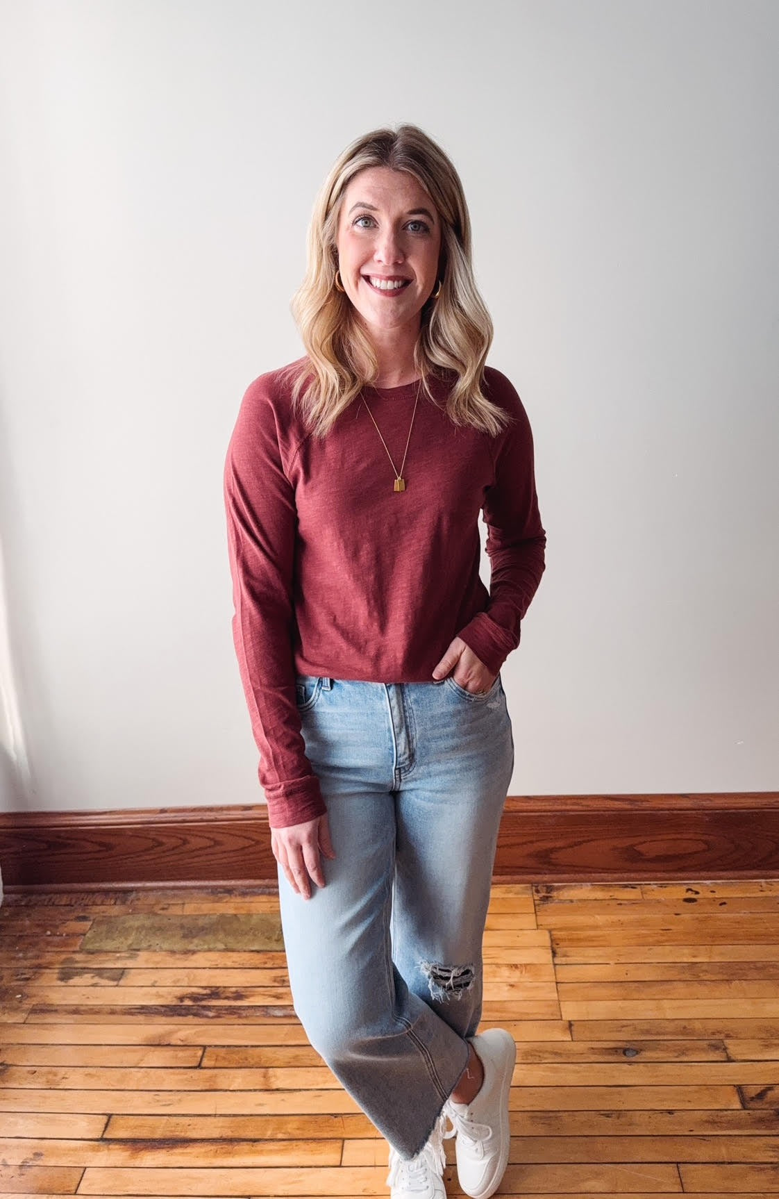 Woman wearing a red sweater and light blue jeans standing on a wooden floor.