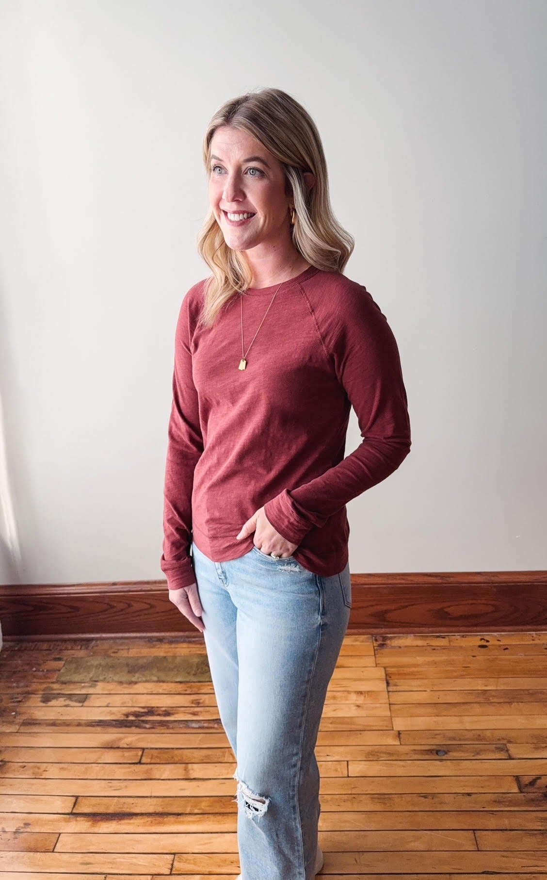 Woman wearing a red sweater and light blue jeans standing on a wooden floor.