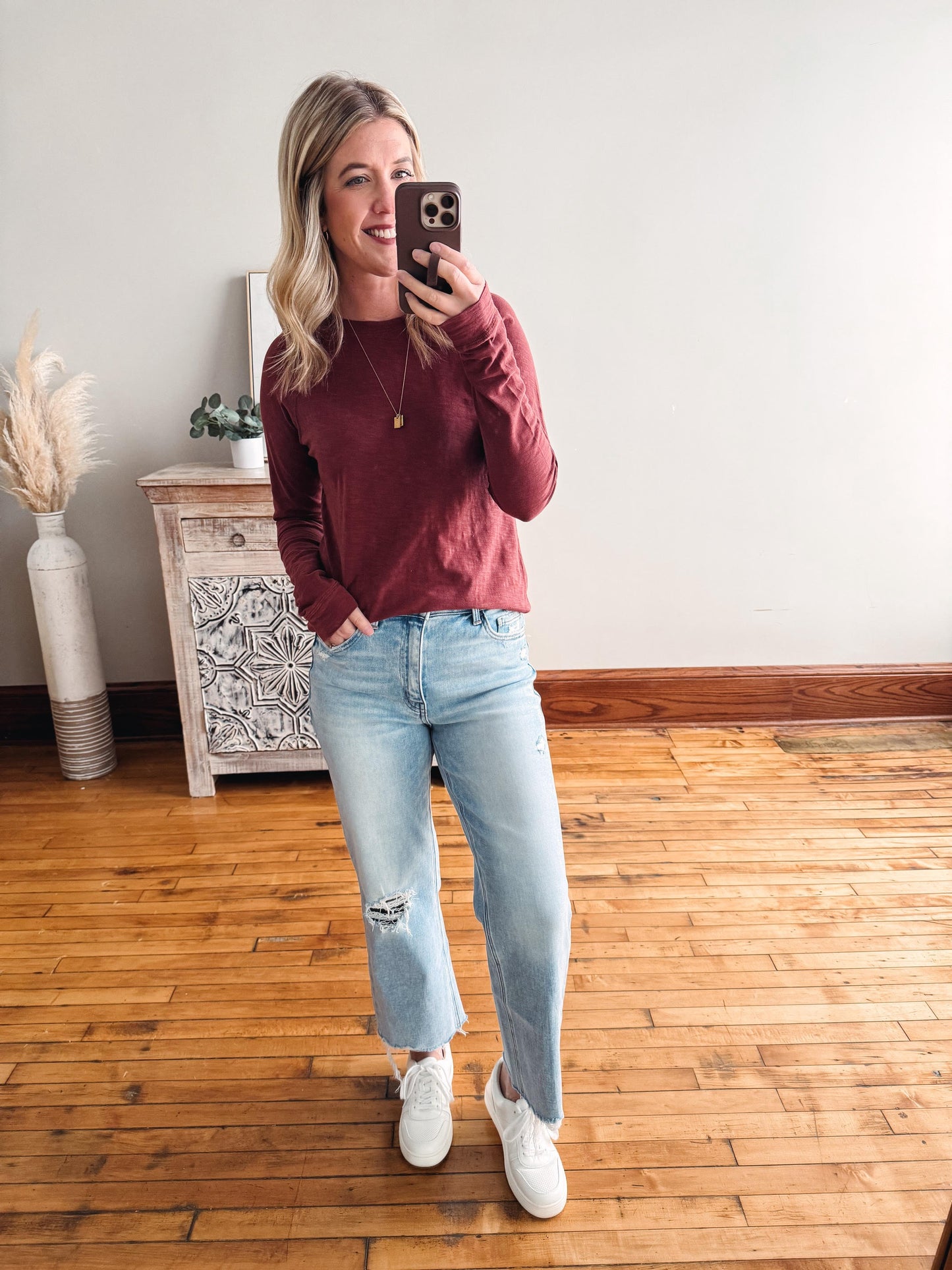 Woman taking a mirror selfie wearing a maroon sweater, light blue jeans, and white sneakers in a room with wooden flooring.