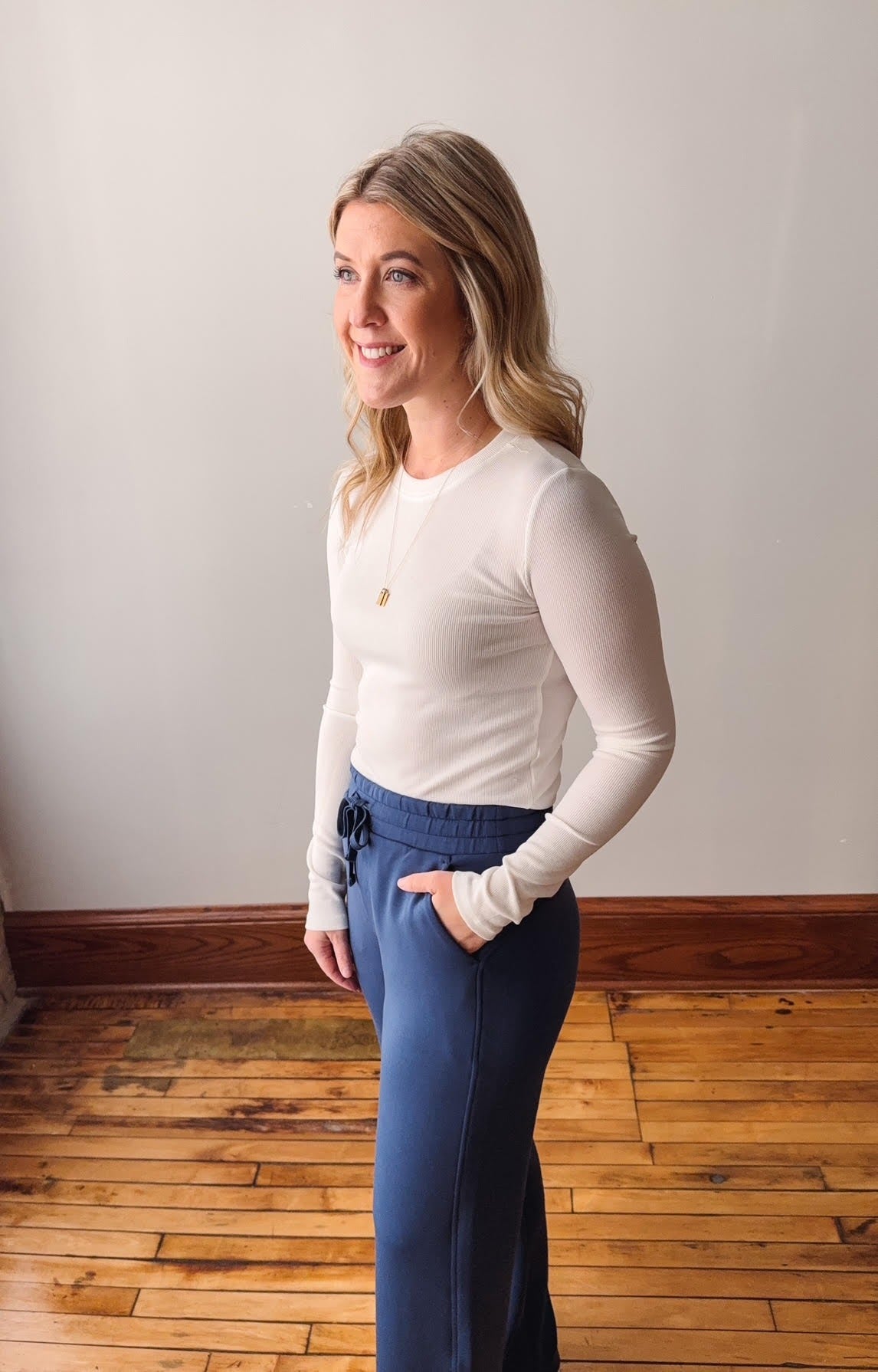 Woman wearing a white long-sleeve top and blue pants standing in a room with wooden flooring.