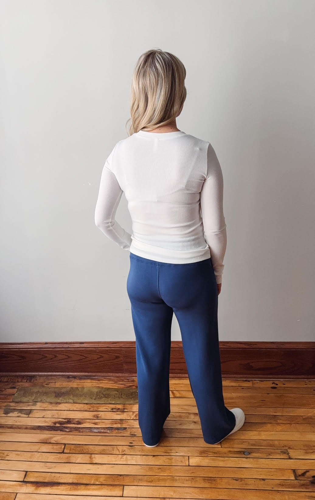 Person wearing a white long-sleeve top and blue pants standing on a wooden floor with a plain wall background.
