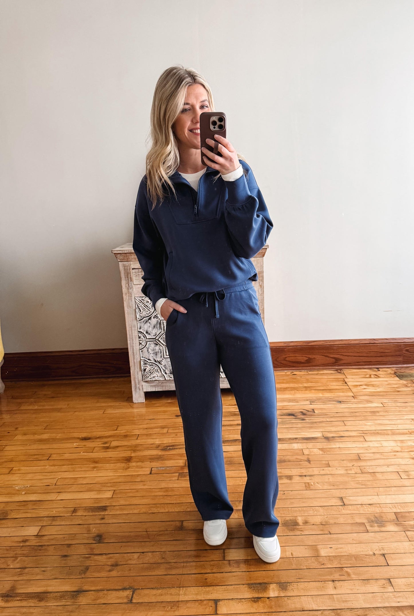 Woman taking a mirror selfie wearing a blue tracksuit in a room with wooden flooring.