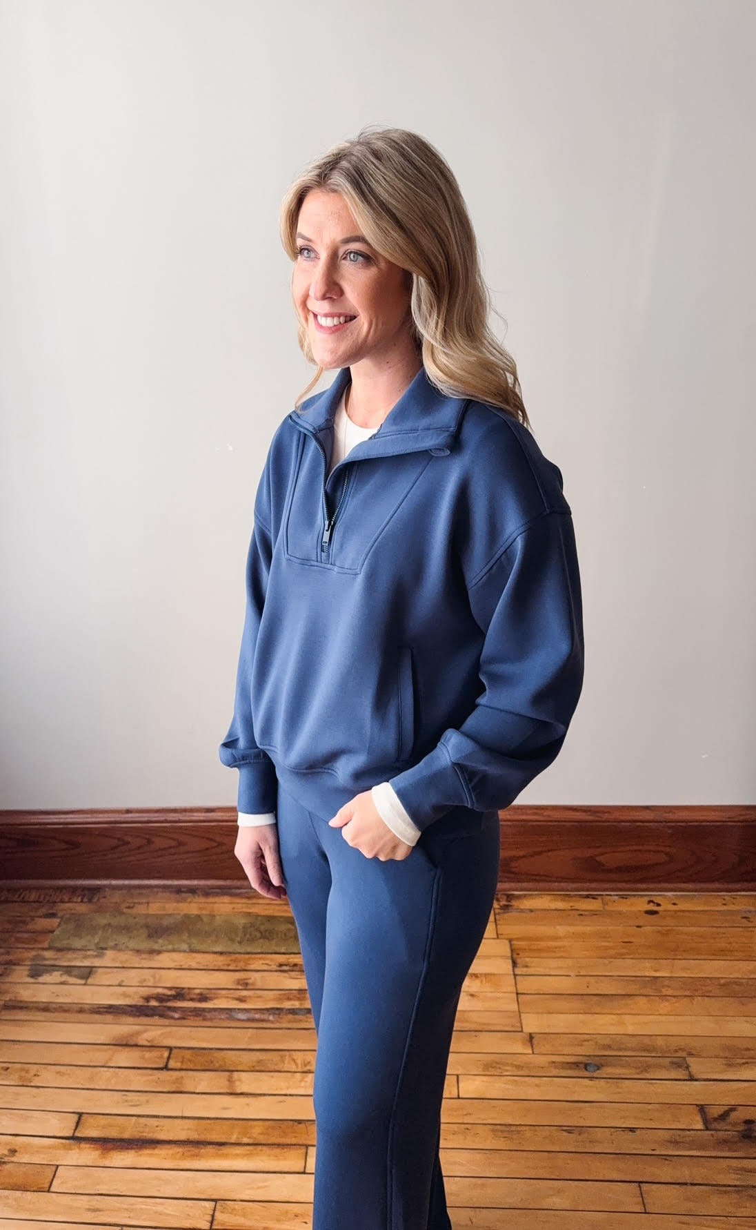 Woman wearing a blue pullover and pants standing on a wooden floor with a white wall background