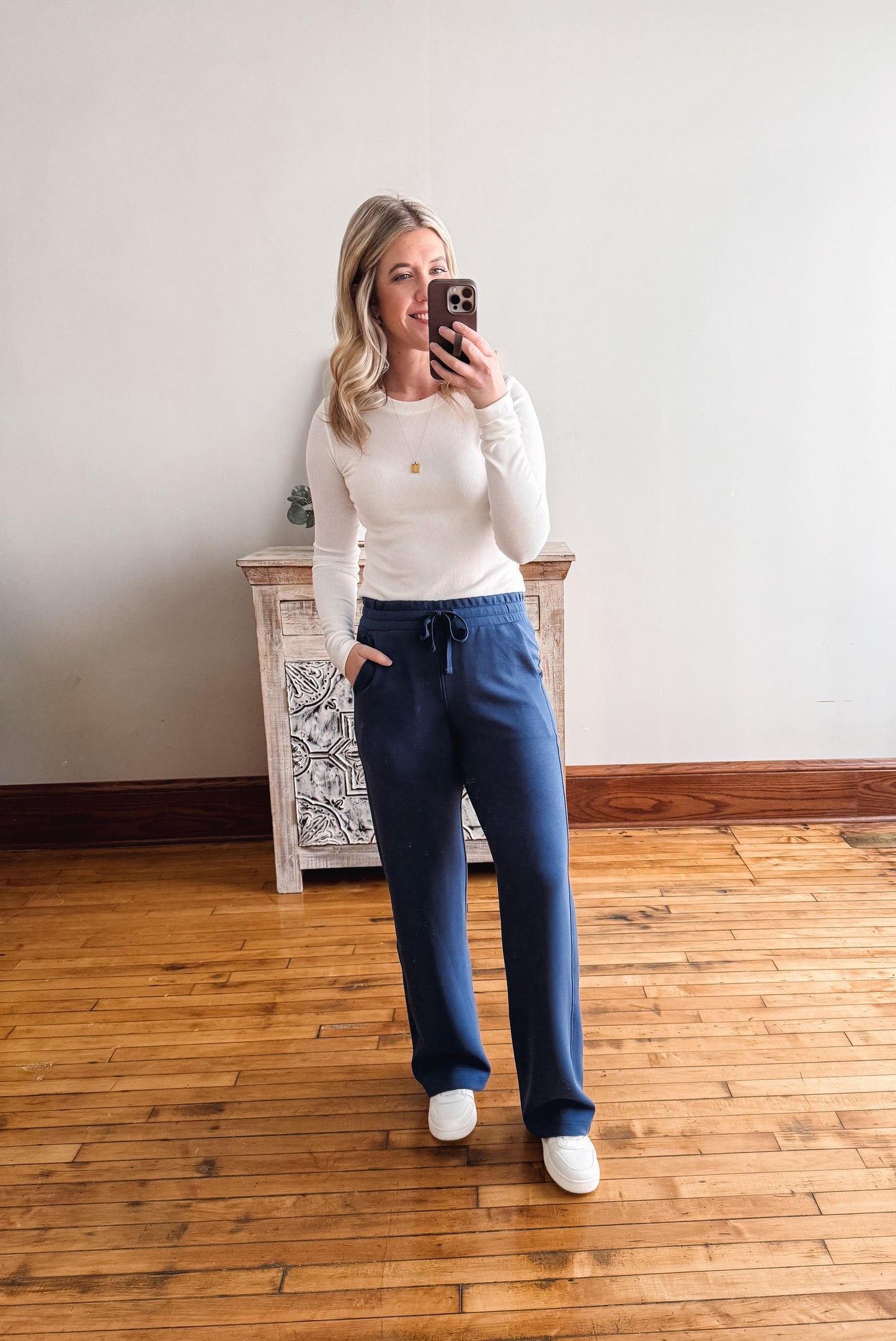 Woman taking a mirror selfie wearing a white long-sleeve top and blue sweatpants on a wooden floor.