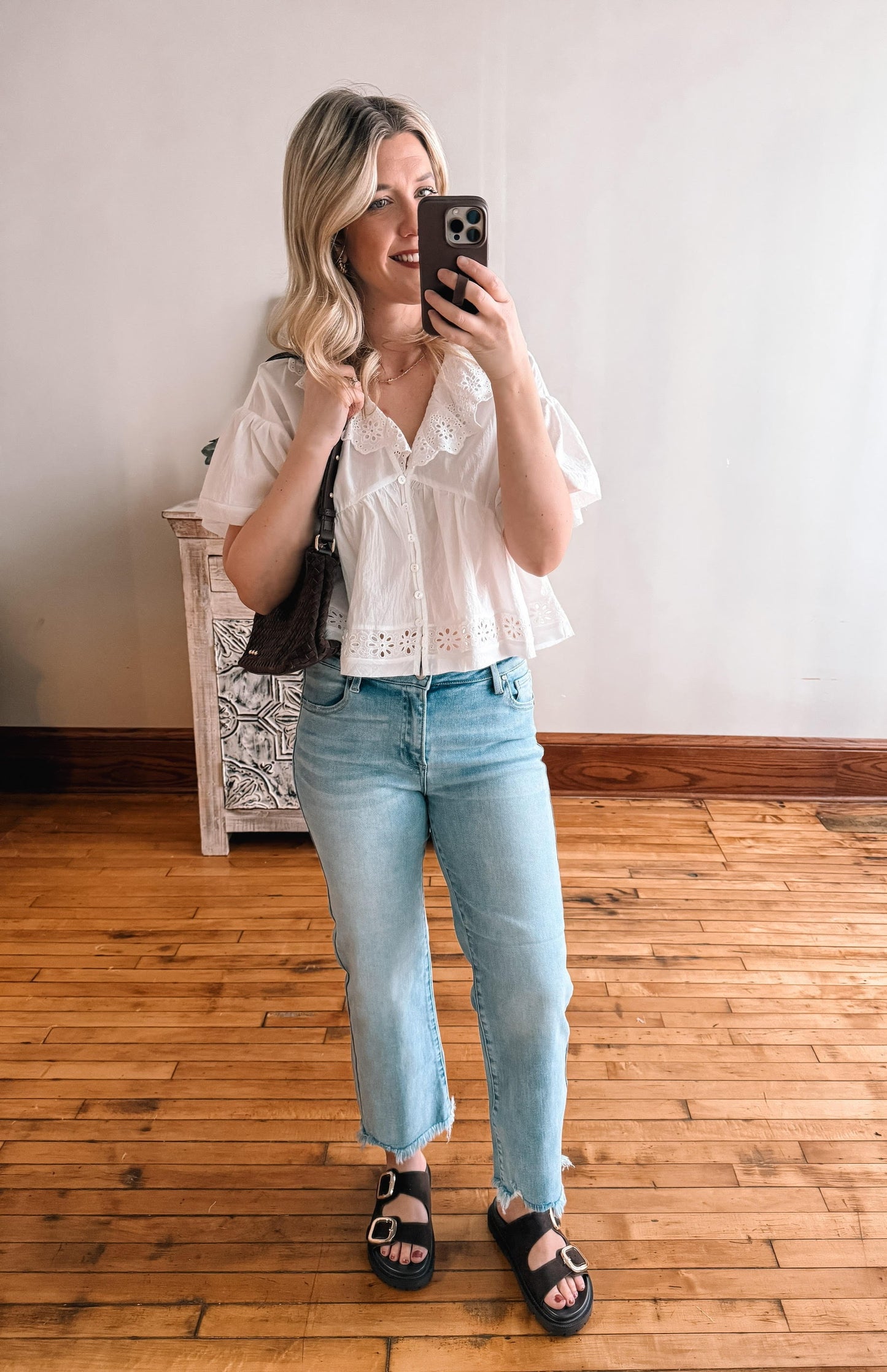 Woman taking a mirror selfie wearing a white blouse and light blue jeans in a room with wooden flooring.
