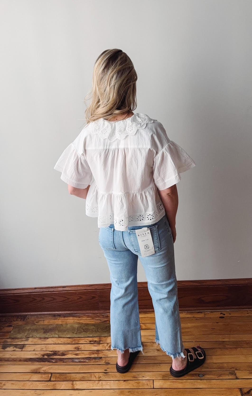 Person wearing a white blouse and light blue jeans with a visible tag, standing against a plain wall.