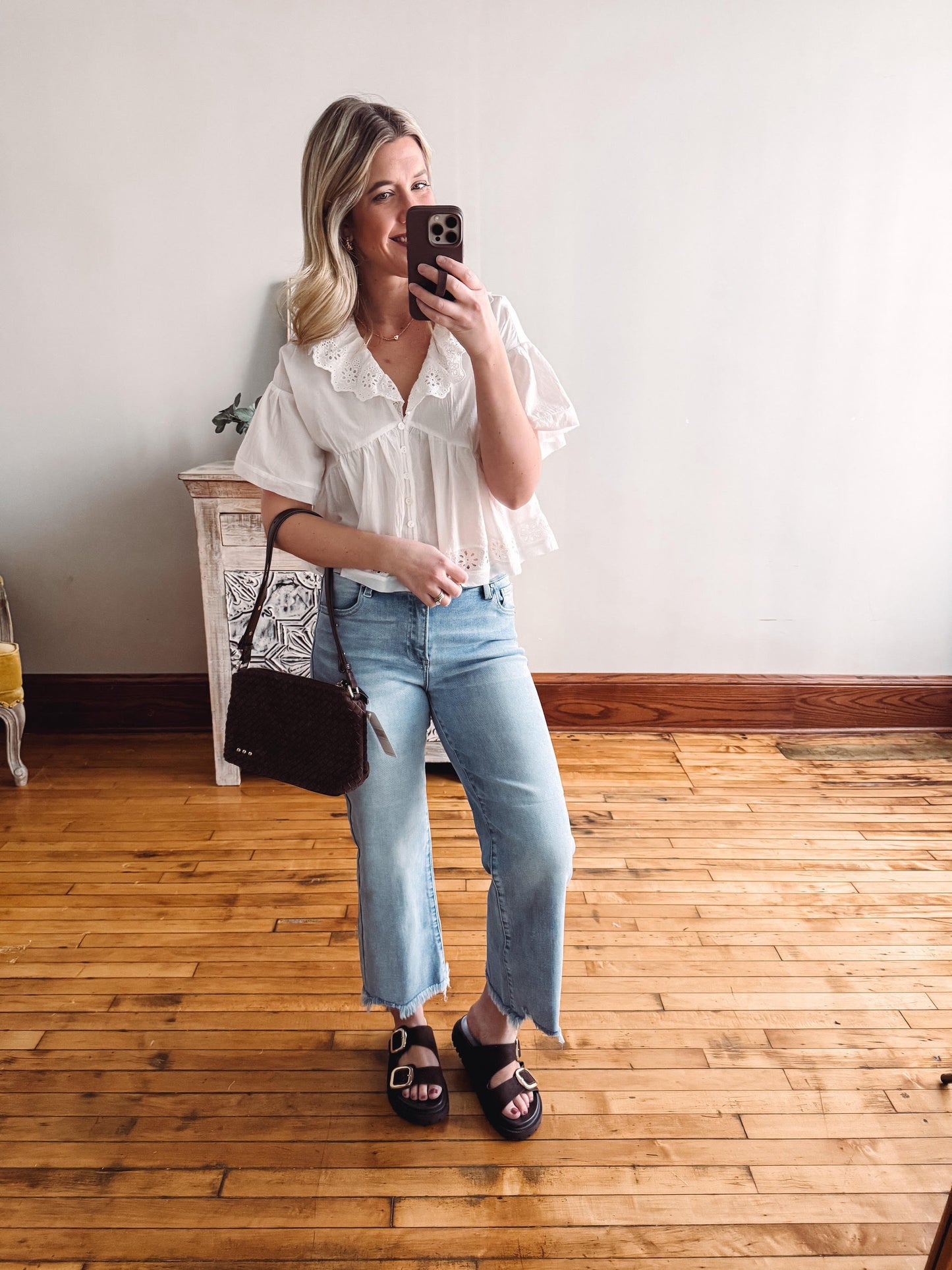 Woman taking a mirror selfie wearing a white blouse and blue jeans in a room with wooden flooring.