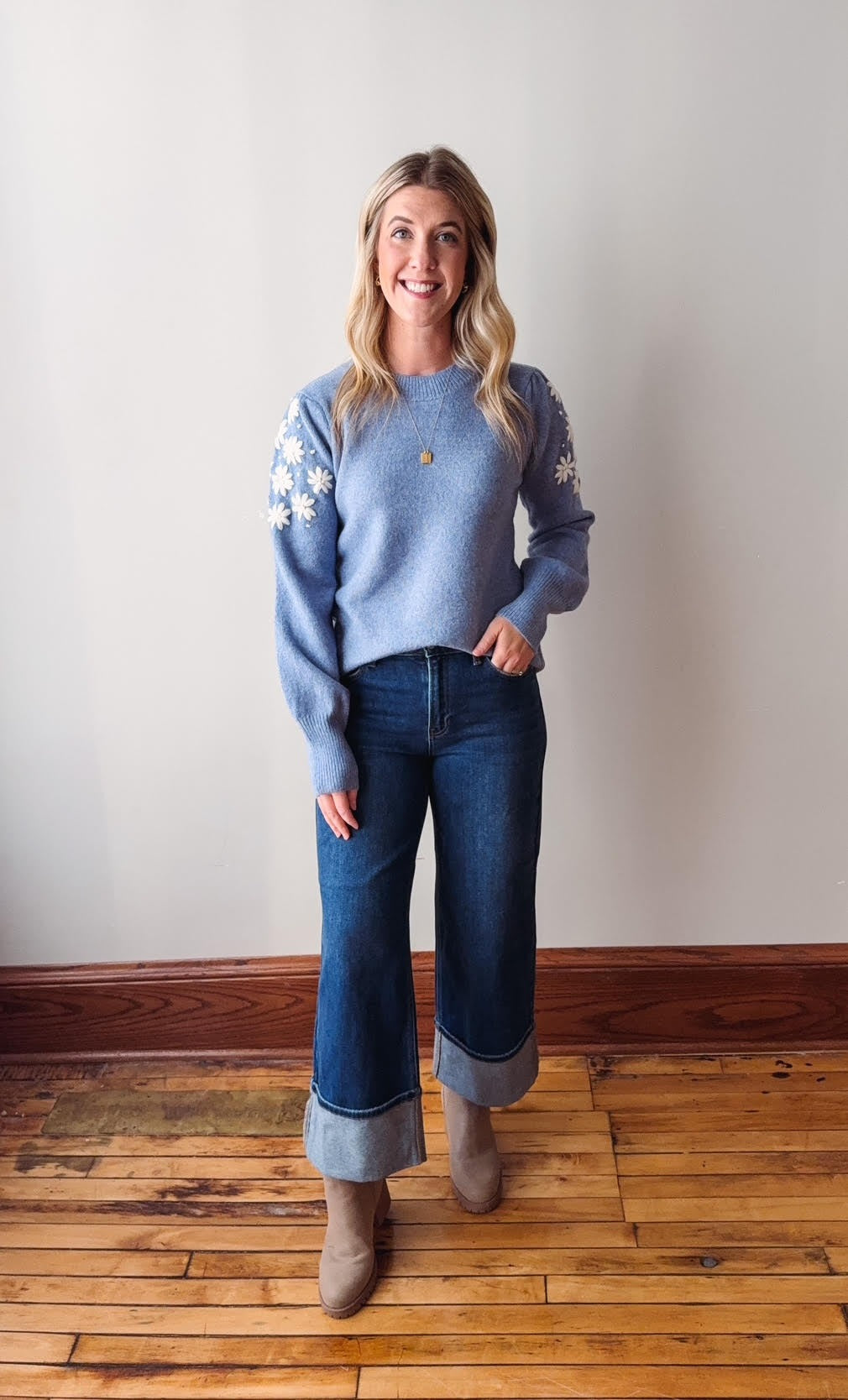 Woman wearing a light blue sweater with floral patterns and blue jeans standing on a wooden floor.