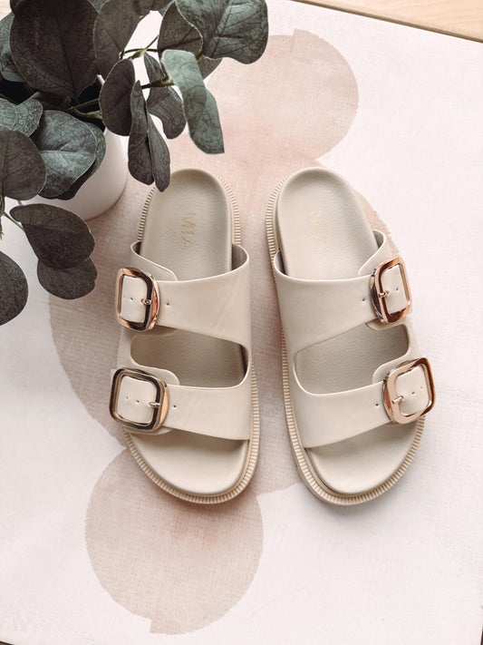 Pair of beige sandals with rose gold buckles on a light surface with eucalyptus leaves.