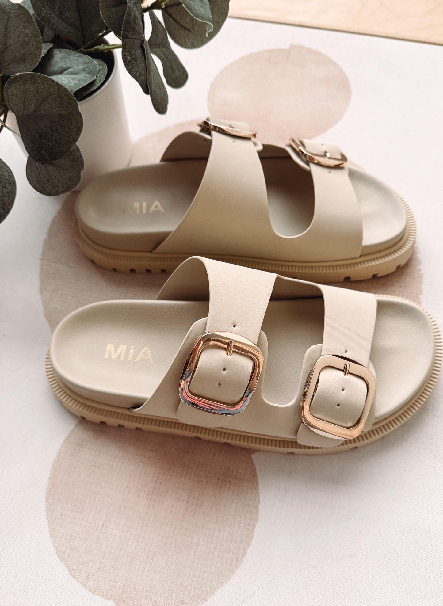 Beige sandals with gold buckles on a white surface with greenery.