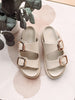 Pair of beige sandals with rose gold buckles on a light surface with eucalyptus leaves.