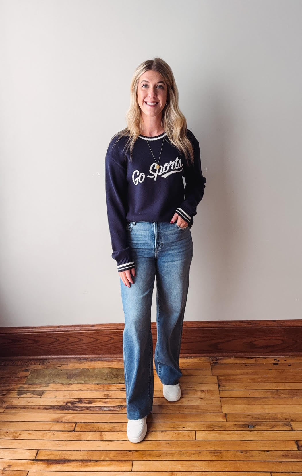 Woman wearing a navy blue sweatshirt with 'Go Sports' text and blue jeans standing on a wooden floor.