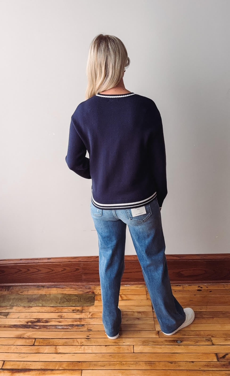 Person wearing a navy sweater and blue jeans standing on a wooden floor with a white wall background