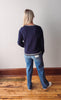 Person wearing a navy sweater and blue jeans standing on a wooden floor with a white wall background