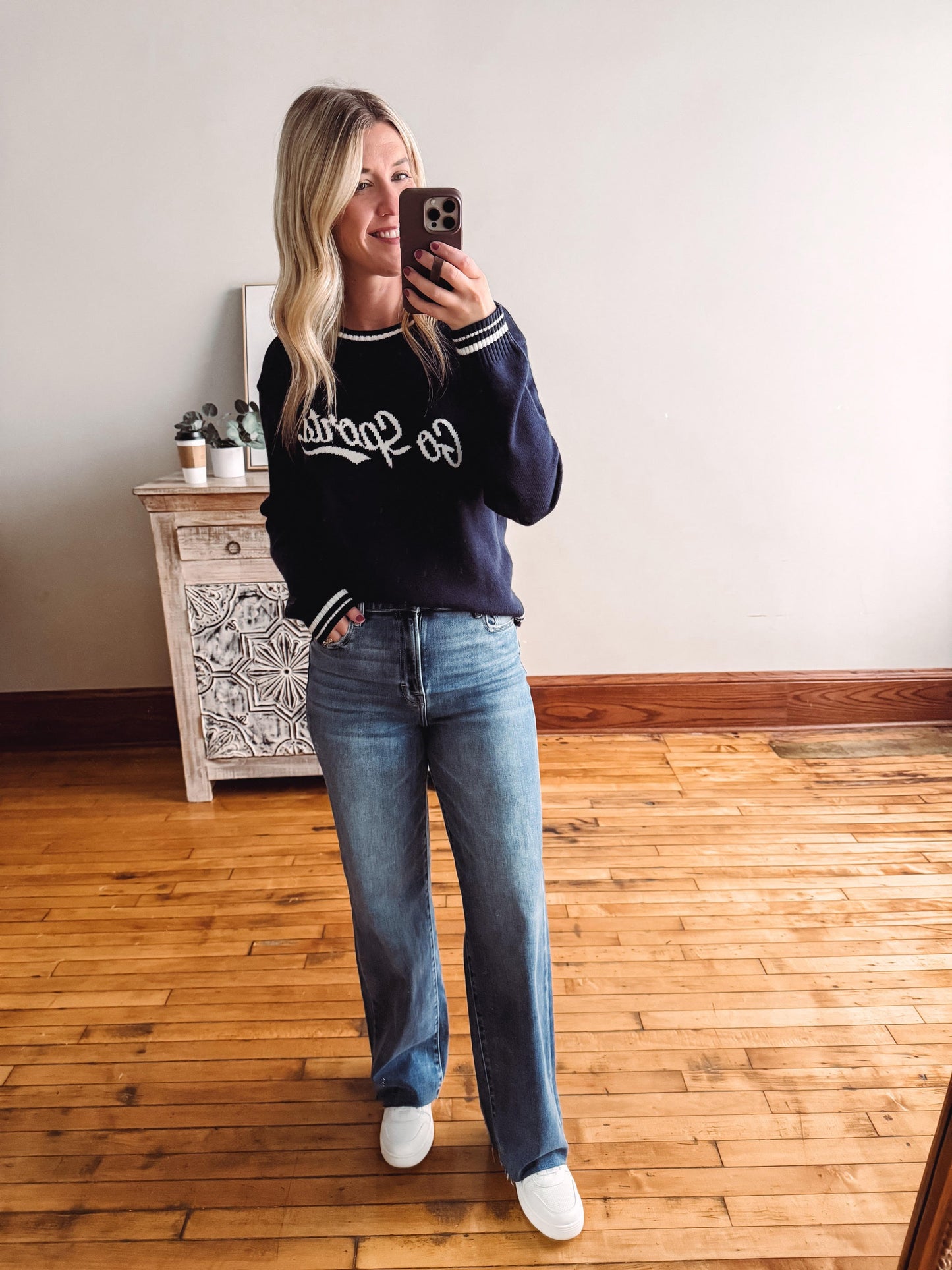 Woman taking a mirror selfie wearing a navy sweater with white lettering and blue jeans on a wooden floor.