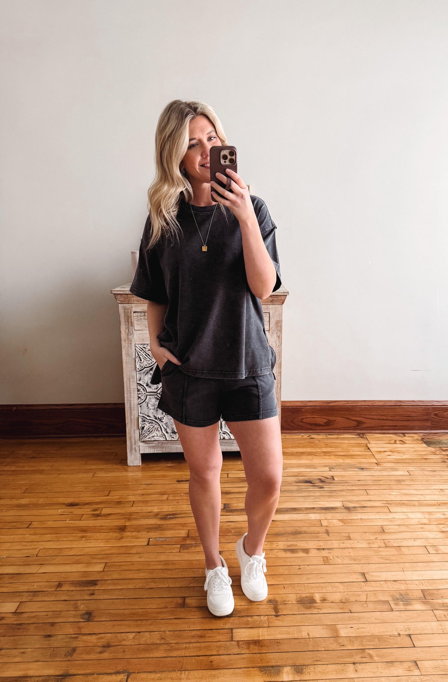 Woman taking a mirror selfie wearing a black t-shirt and shorts on a wooden floor.