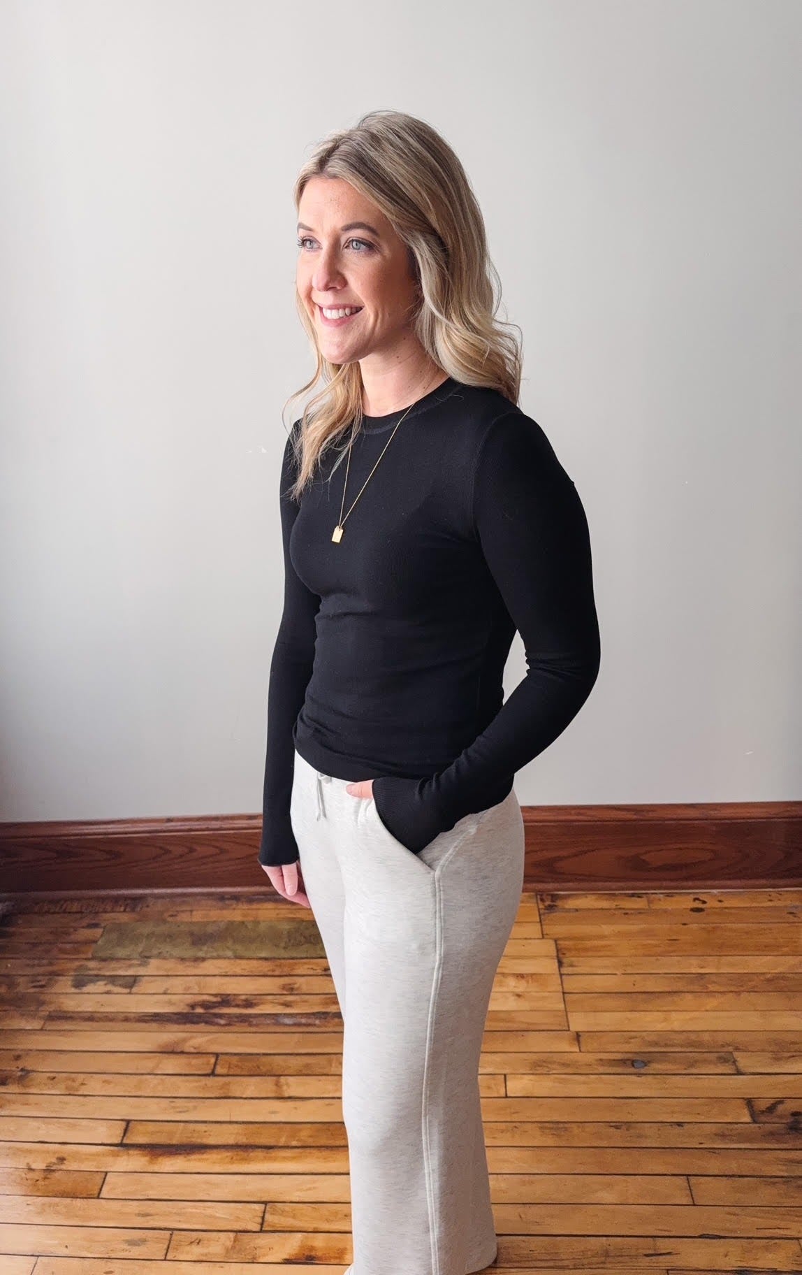 Woman wearing a black long-sleeve top and white pants standing on a wooden floor.