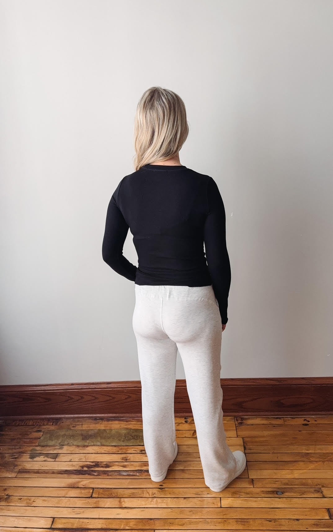 Person wearing a black top and white pants standing against a plain wall.