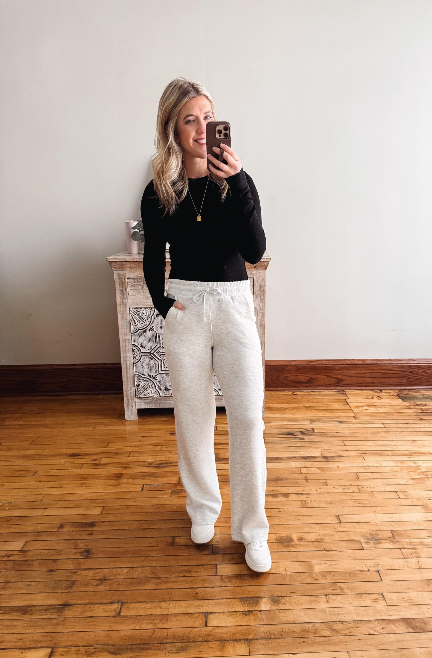 Woman taking a mirror selfie wearing a black top and white pants in a room with wooden flooring and a decorative cabinet.