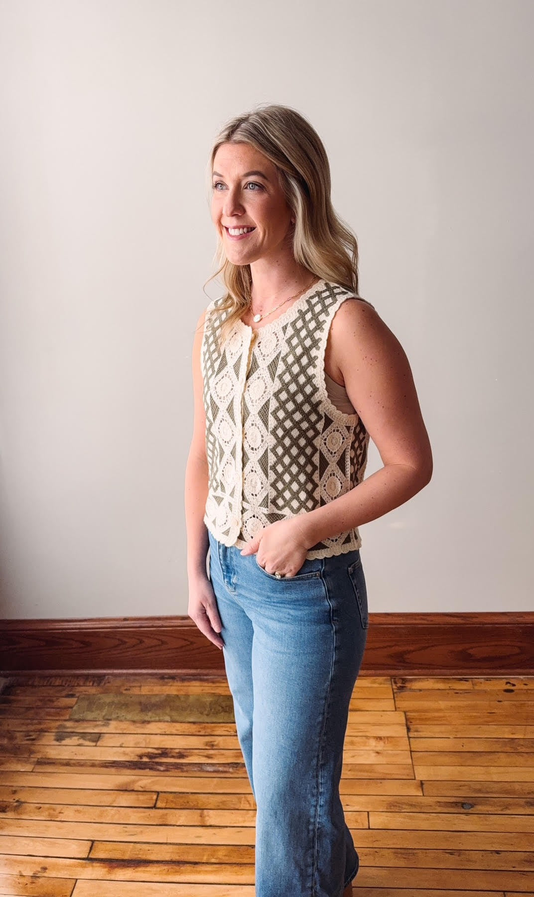 Woman wearing a sleeveless patterned top and blue jeans standing in a room with wooden flooring.