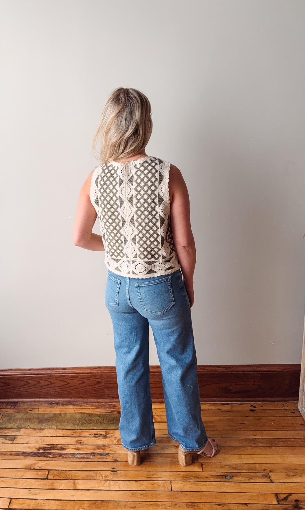 Person wearing a sleeveless patterned top and blue jeans standing on a wooden floor.