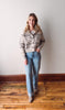 Woman wearing a patterned sweater and jeans standing in a room with wooden flooring and a white wall.