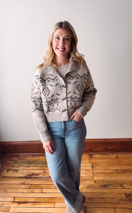Woman wearing a patterned cardigan and jeans standing on a wooden floor.