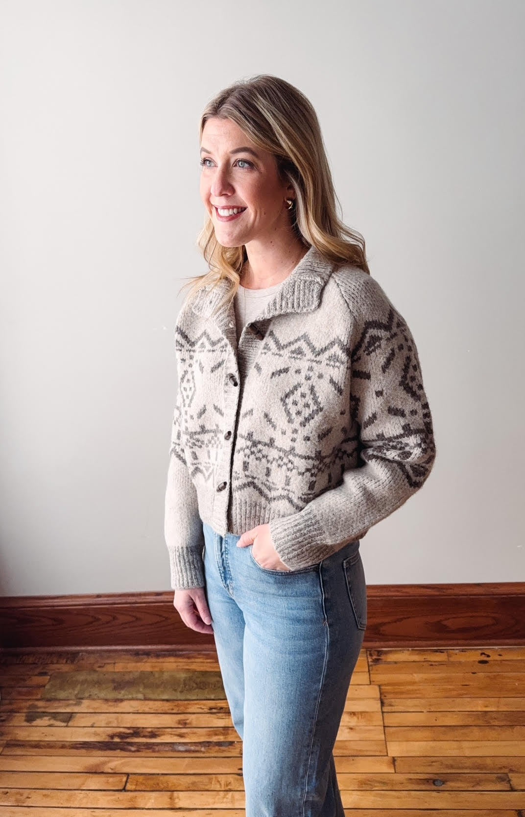 Woman wearing a patterned cardigan and jeans against a plain wall.
