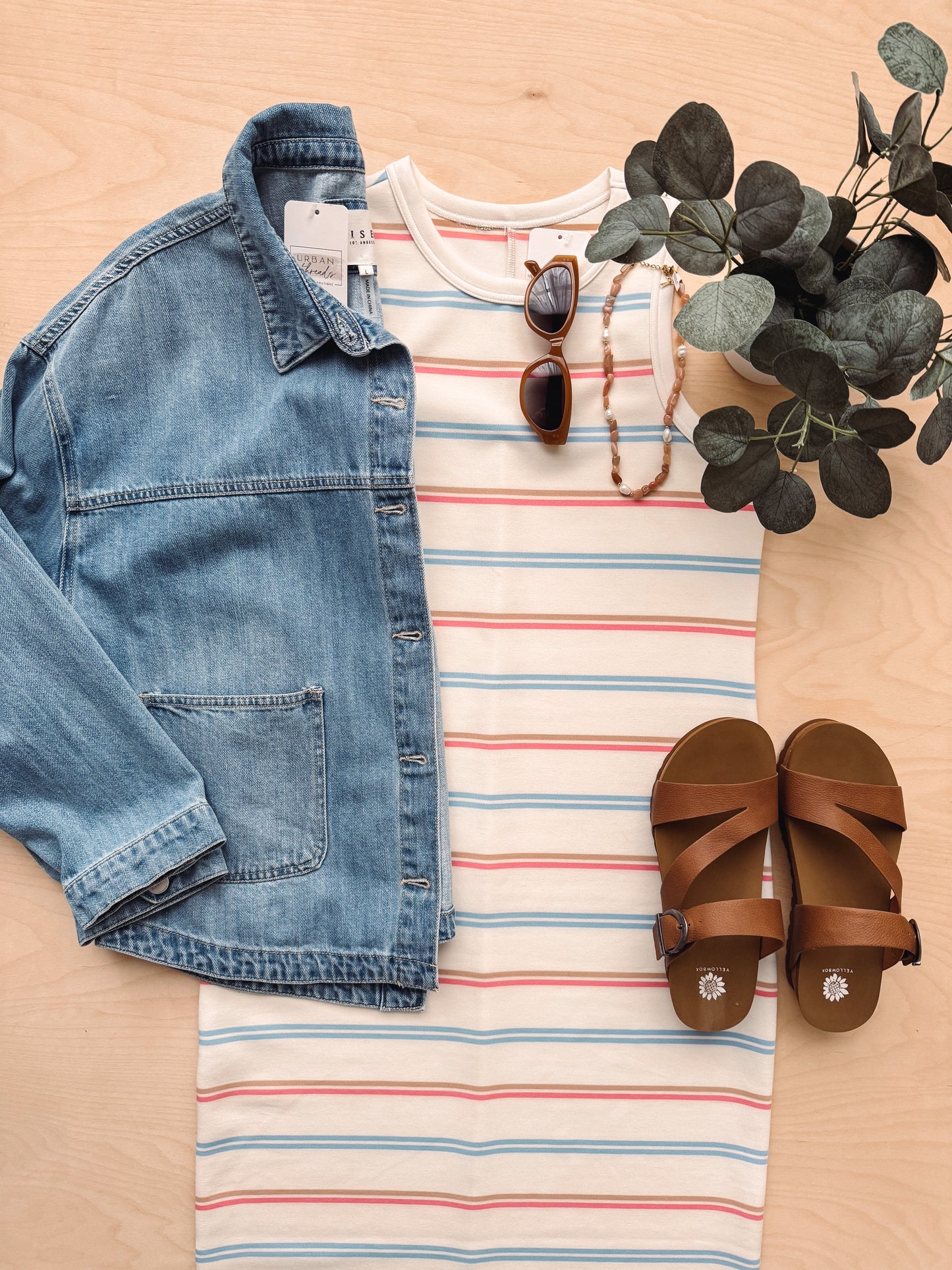 Denim jacket, striped shirt, sunglasses, necklace, and sandals on a wooden surface with eucalyptus leaves.