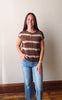 A young woman stands indoors wearing a brown and beige horizontally striped short-sleeve zip-up knit top with blue jeans.