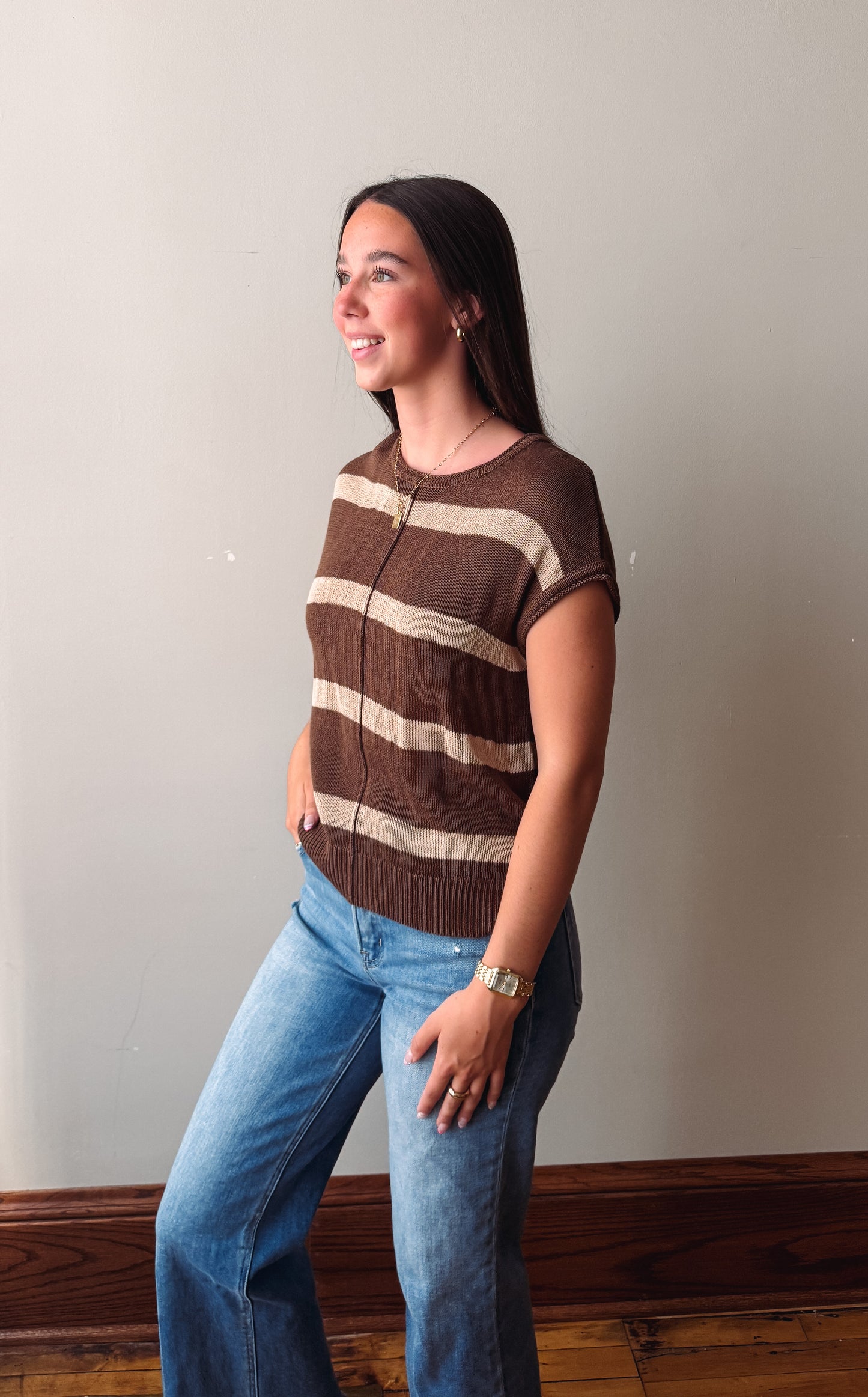 A woman wearing a brown and beige horizontally striped short-sleeve knit top and blue jeans, standing against a plain light gray wall.