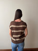 A woman viewed from behind wearing a brown and beige horizontally striped short-sleeve knit top and blue jeans, standing against a plain light gray wall.