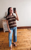 A woman takes a mirror selfie in a room with hardwood floors and a white wall, wearing a brown and beige horizontally striped knit top, blue cropped jeans, and brown leather slide sandals.