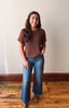 A woman modeling a brown short-sleeve knit top and blue wide-leg jeans, standing on a wooden floor against a plain off-white wall.