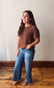 A woman modeling a brown short-sleeve knit top and blue wide-leg jeans, standing on a wooden floor against a plain off-white wall.