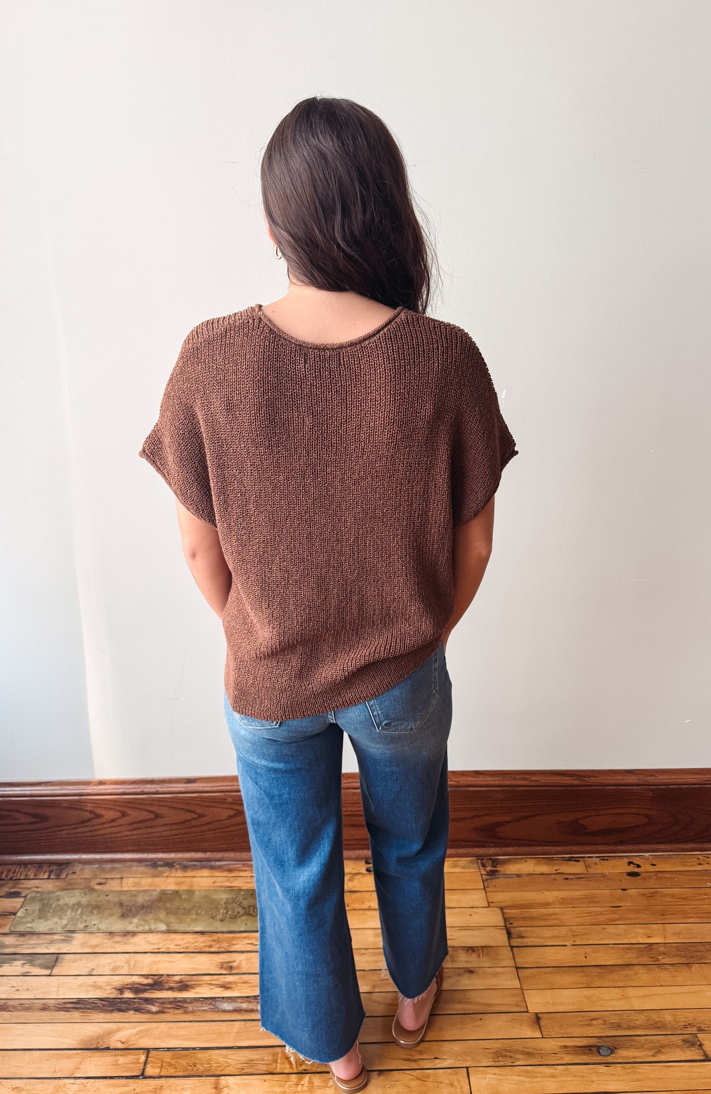 A woman viewed from behind wearing a brown short-sleeve knit top and blue jeans, standing on a wooden floor against a plain white wall.
