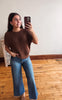 A woman taking a mirror selfie in an indoor room with hardwood floors and a white wall, wearing a brown short-sleeve knit sweater and blue wide-leg jeans.