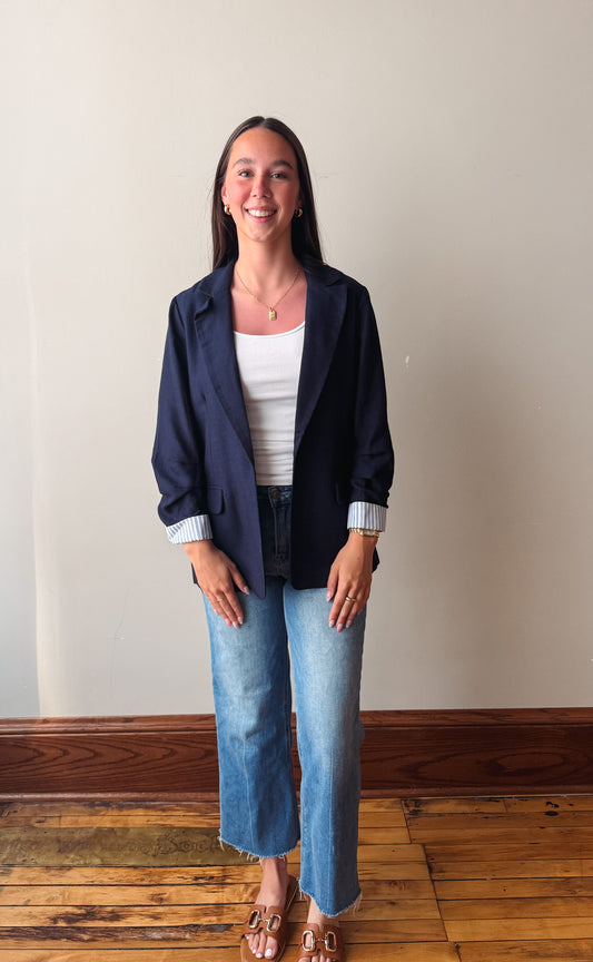 A young woman stands in a room with a beige wall and wooden floor, wearing a navy blue blazer over a white ribbed tank top, blue jeans, and brown flat sandals.
