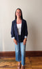 A young woman stands in a room with a beige wall and wooden floor, wearing a navy blue blazer over a white ribbed tank top, blue jeans, and brown flat sandals.