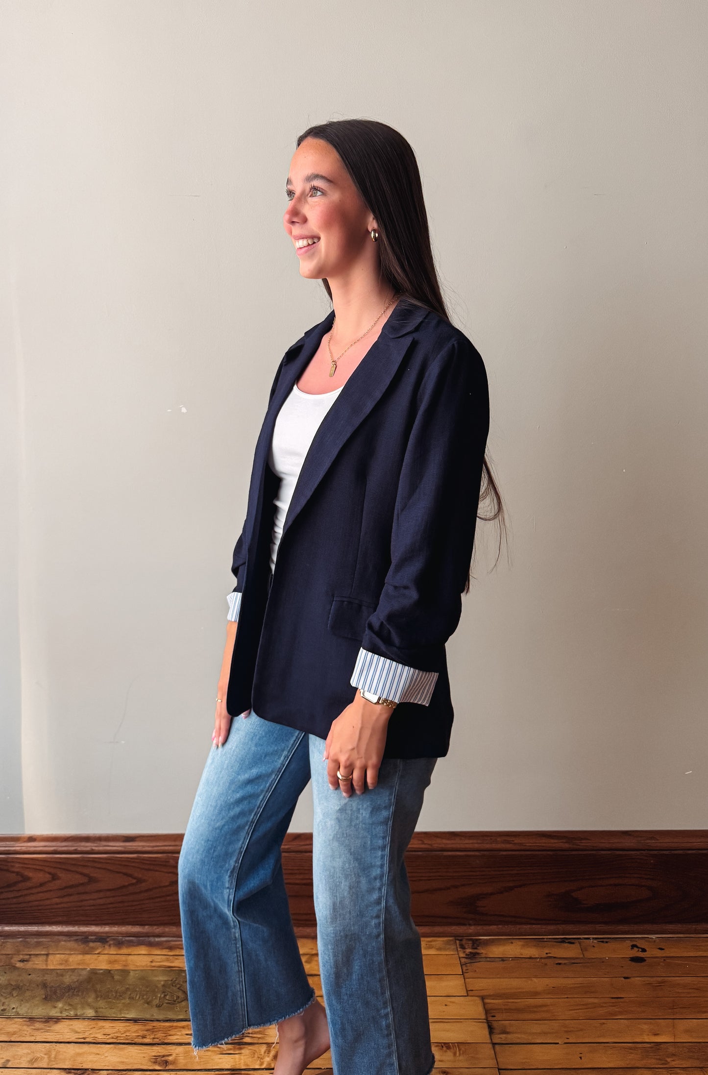 A woman wearing a navy blue blazer over a white top and blue jeans, standing in profile against a plain beige wall with a wooden floor.
