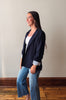 A woman wearing a navy blue blazer over a white top and blue jeans, standing in profile against a plain beige wall with a wooden floor.