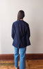 A woman viewed from behind wearing a dark navy blue button-up shirt and light blue jeans, standing on a wooden floor against a plain off-white wall.