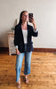 A woman taking a mirror selfie in a room with hardwood floors and a light-colored wall. She is wearing a dark navy blazer over a white tank top, light wash blue jeans, and brown flat sandals.