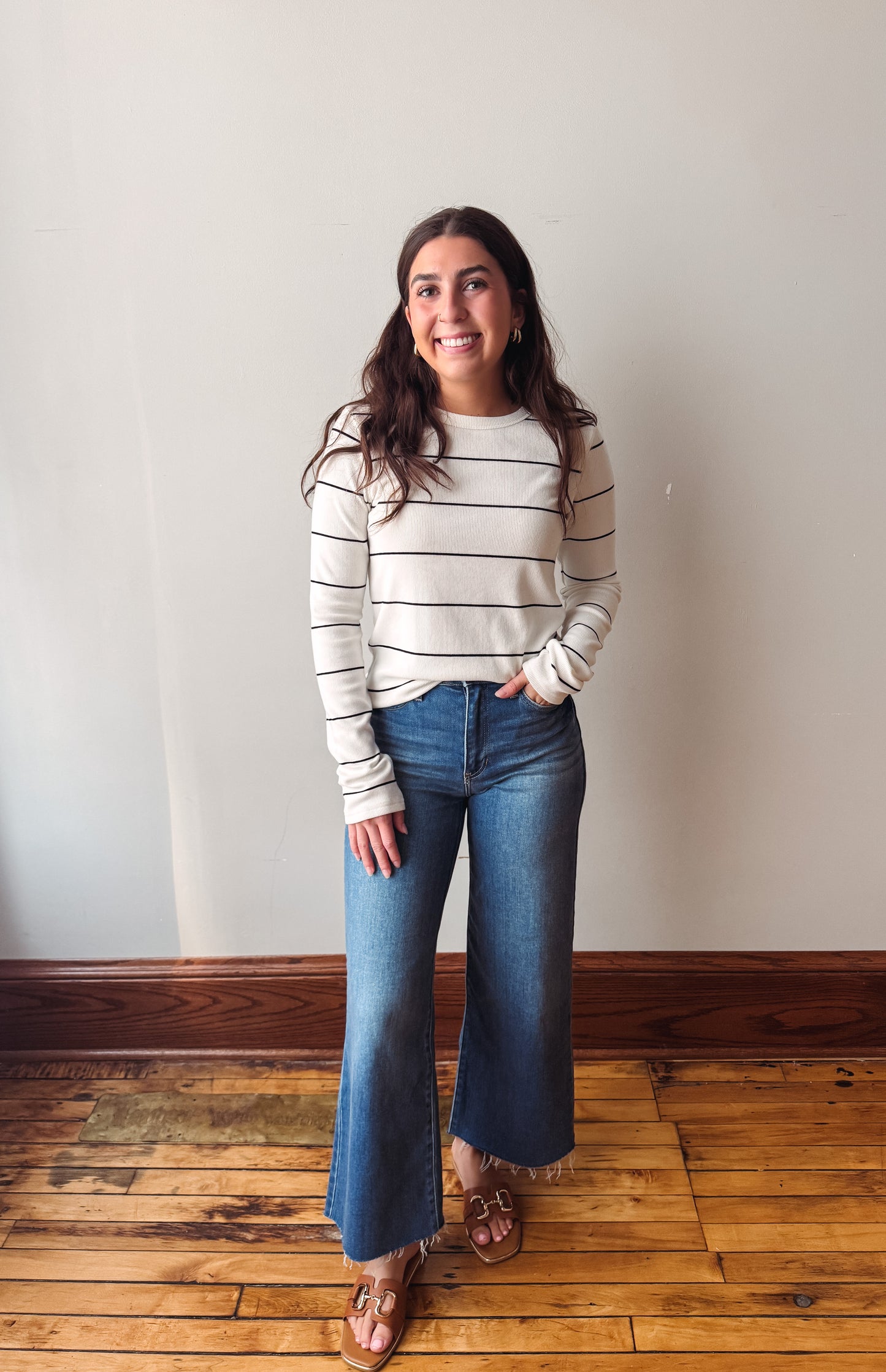 A woman stands in a room with a light beige wall and wooden floor, wearing a cream and black horizontally striped long-sleeve knit sweater, blue wide-leg jeans, and brown leather sandals.