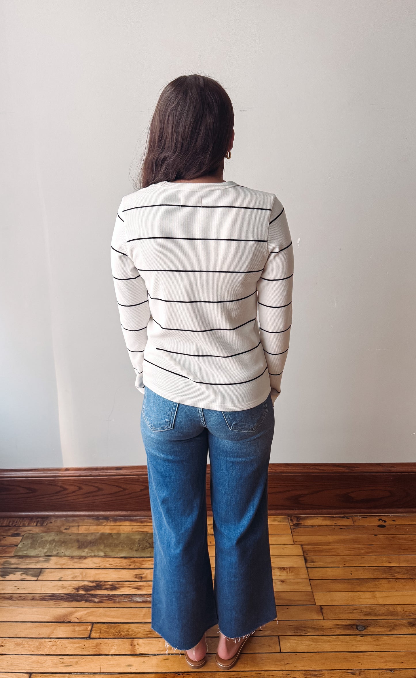 A woman viewed from behind wearing a cream knit sweater with thin black horizontal stripes and blue jeans, standing on a wooden floor against a plain off-white wall.