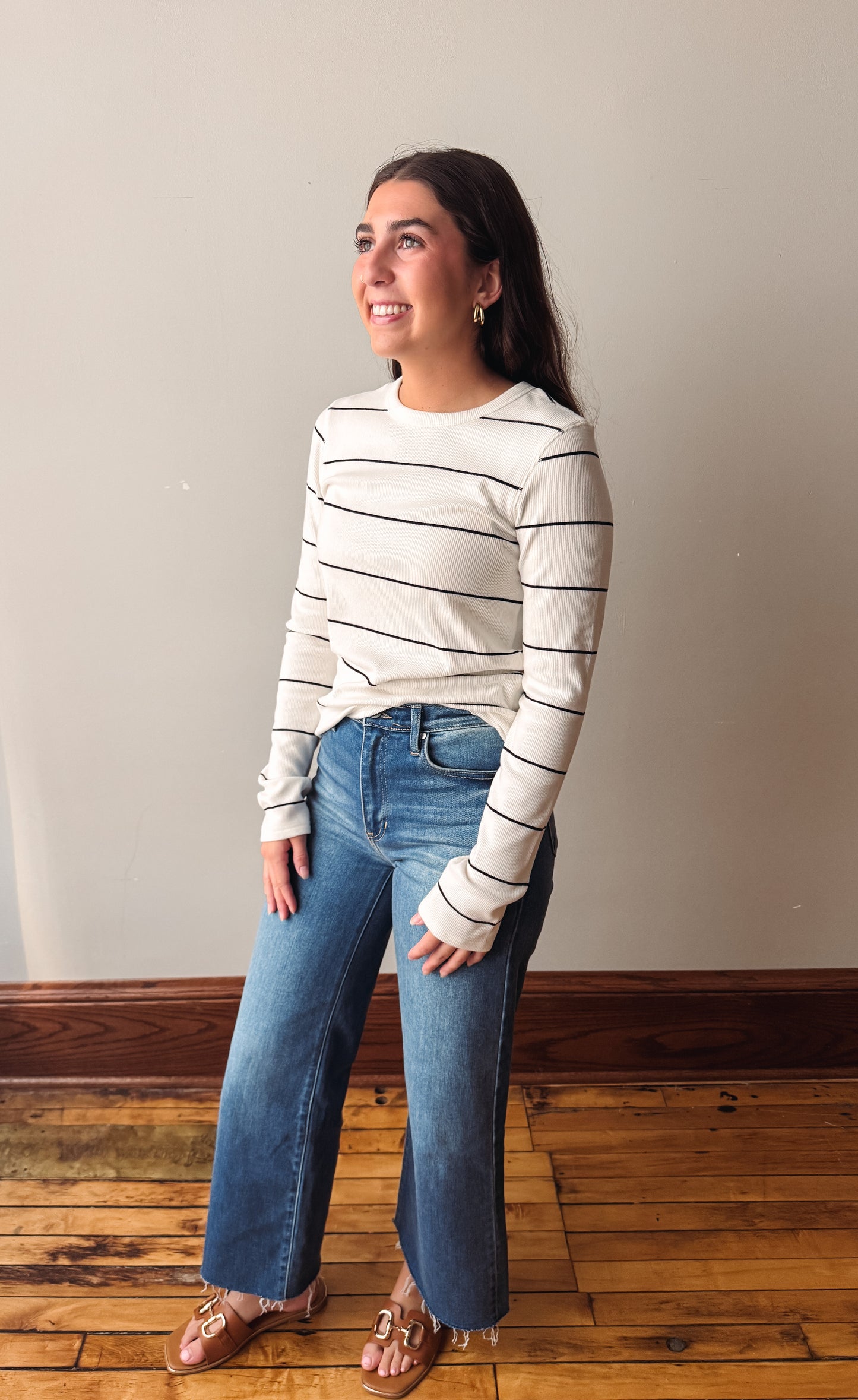 A woman wearing a cream long-sleeve crewneck shirt with thin black horizontal stripes, paired with blue wide-leg jeans and brown leather sandals, standing in a room with a beige wall and wooden floor.