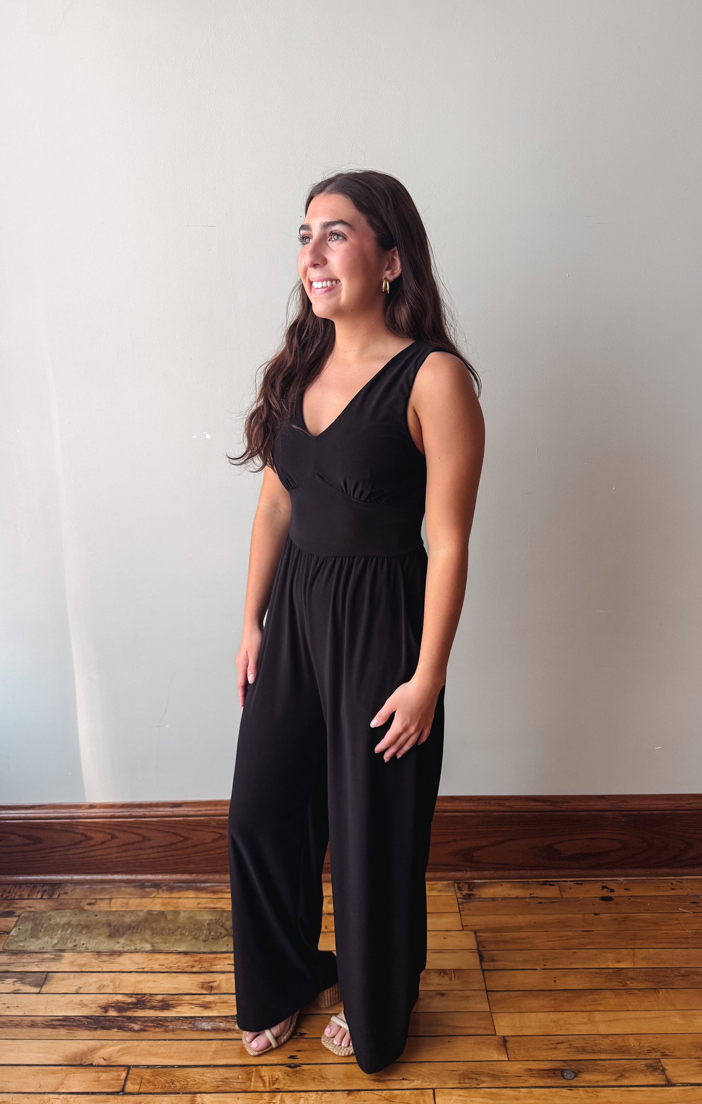 A woman modeling a black sleeveless jumpsuit with a V-neck bodice and wide legs, standing on a wooden floor against a plain off-white wall.